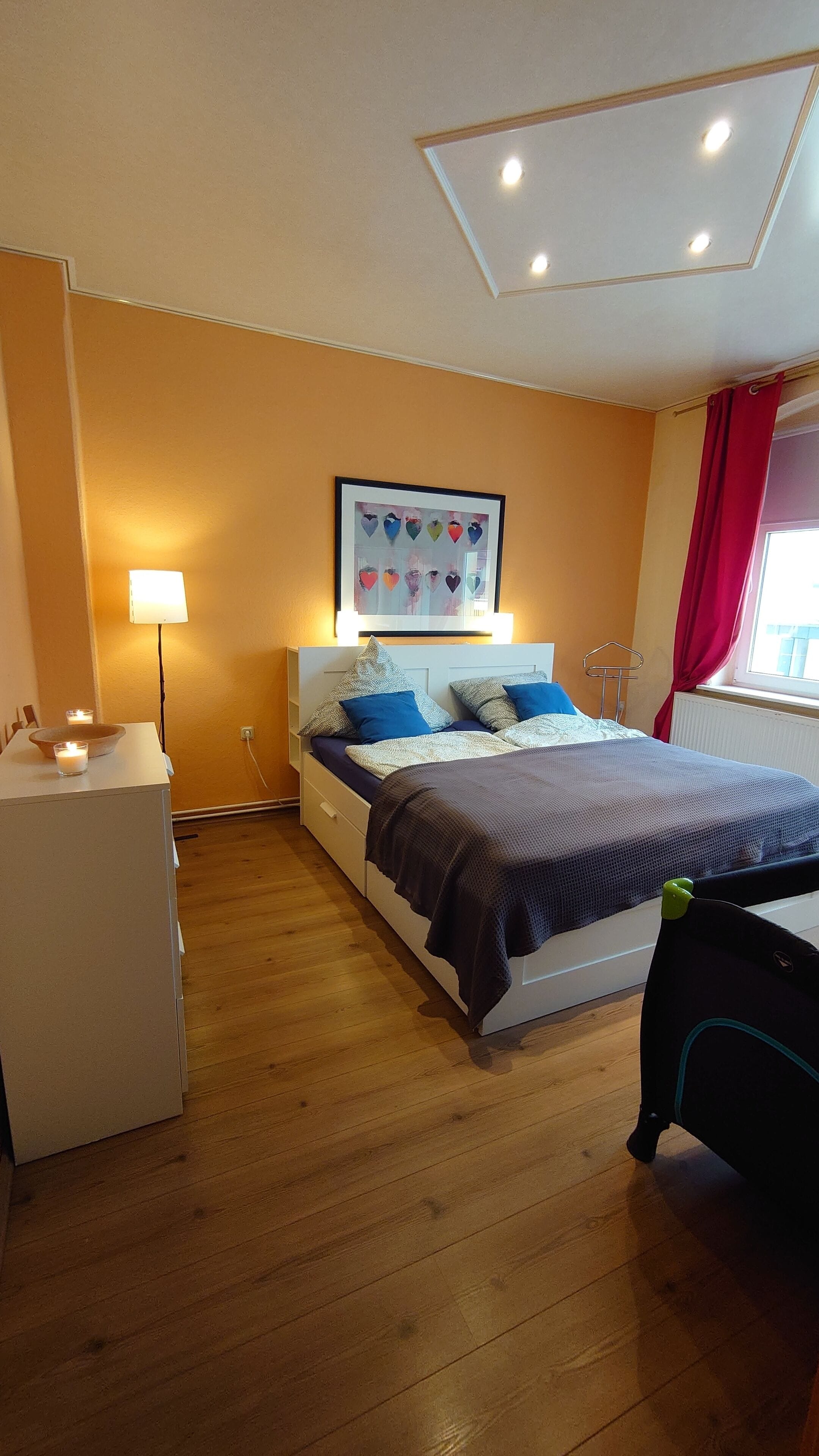 Quarto com cama, mesas de cabeceira, luminária, quadro na parede e janela com cortinas.