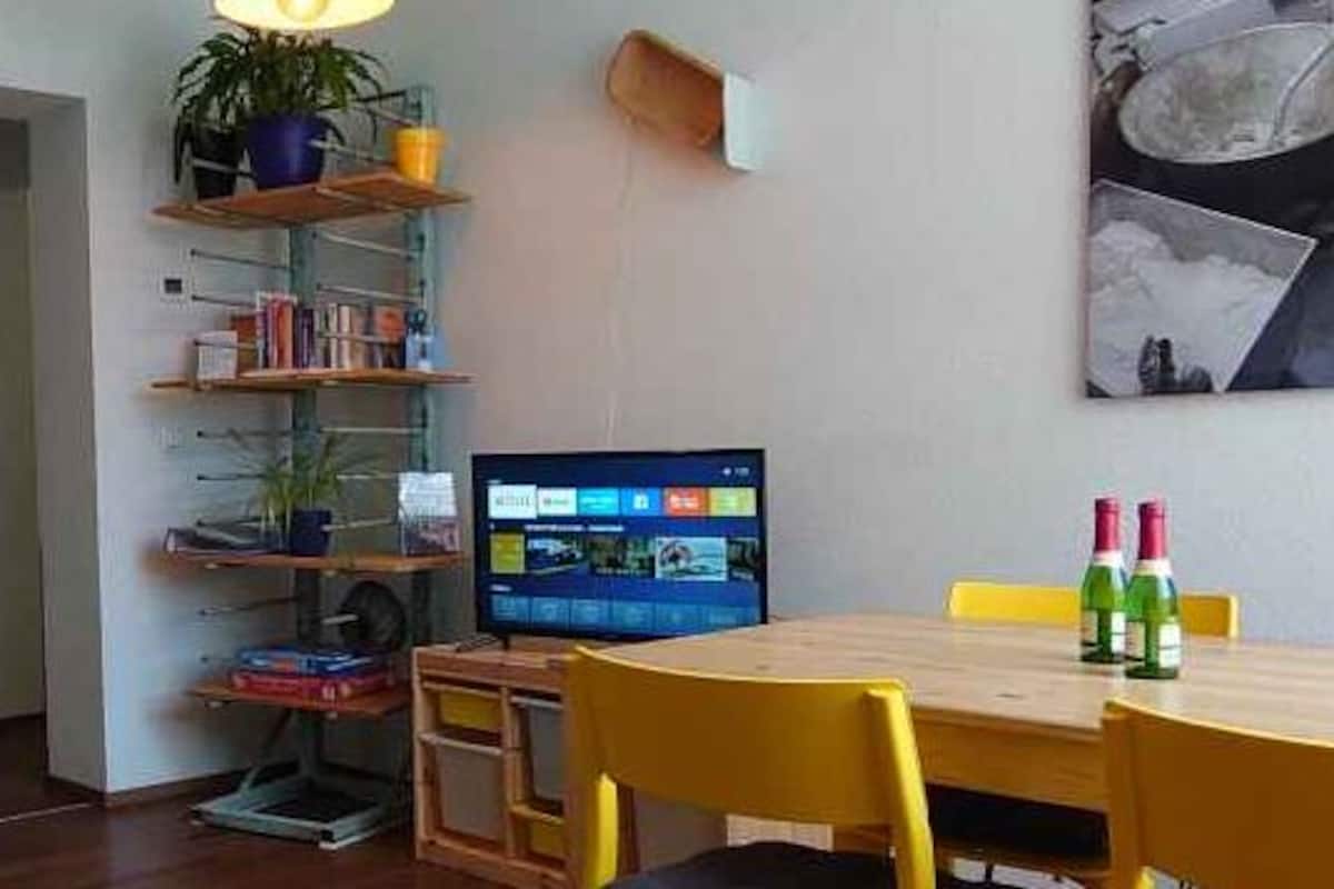 Ein Zimmer mit einem Holztisch, gelben Stühlen, einem blauen Sofa, einem Bücherregal und einem Bild an der Wand.