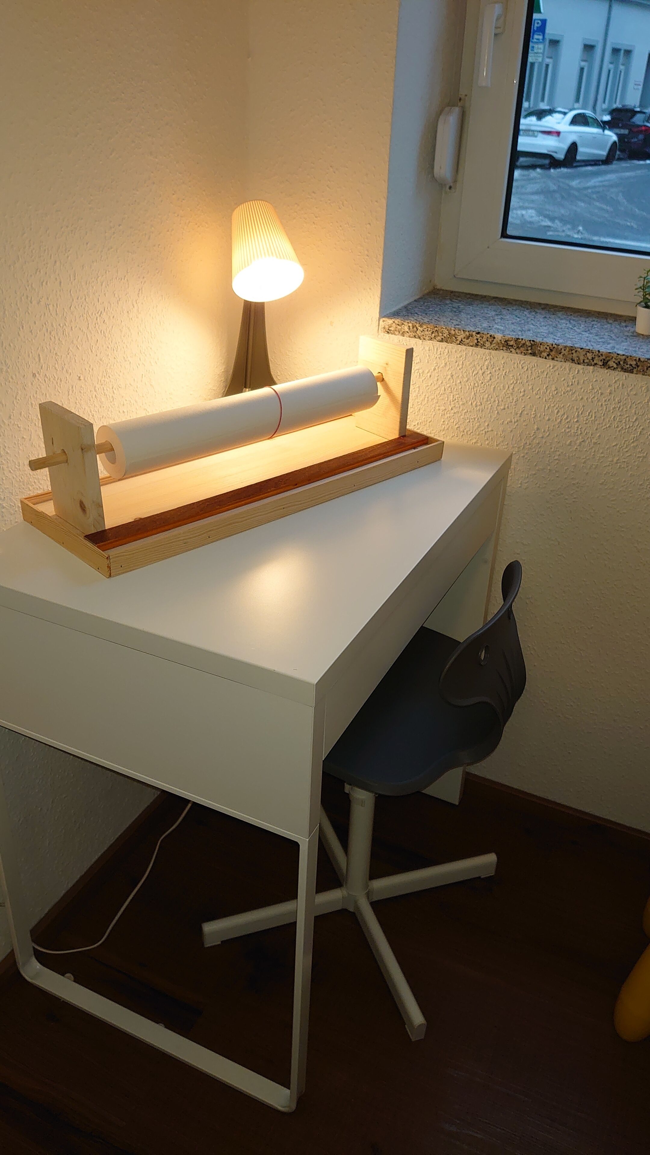 Un escritorio blanco con una lámpara, una silla y una ventana que da a vista de coches estacionados.