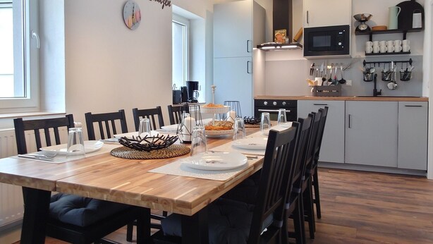 Une cuisine moderne avec une table à manger en bois, des chaises noires, des luminaires suspendus et une horloge fixée au mur.