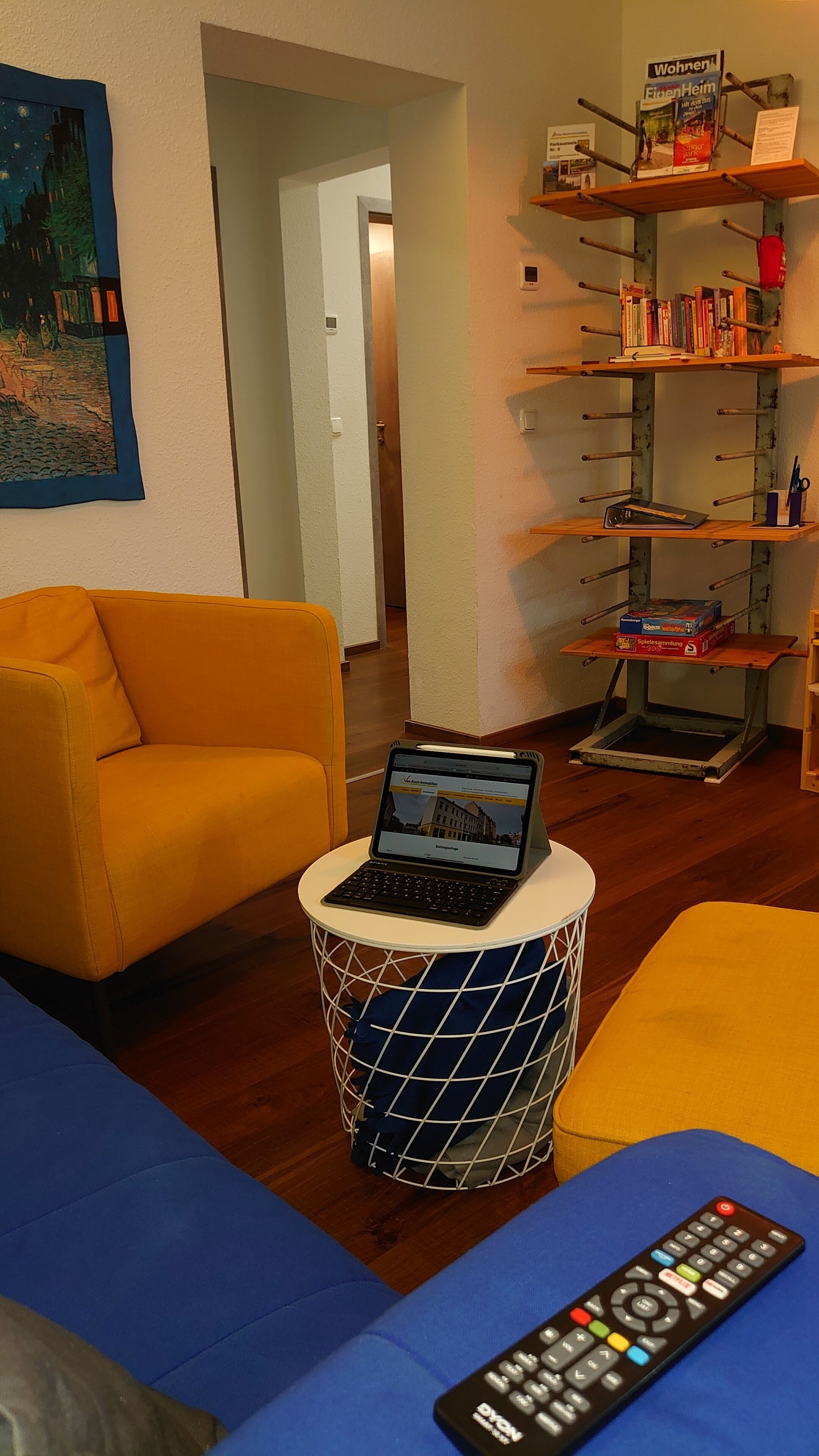 A room with a yellow sofa, a laptop on a wire basket, a remote control, and a bookshelf with books and a sign.