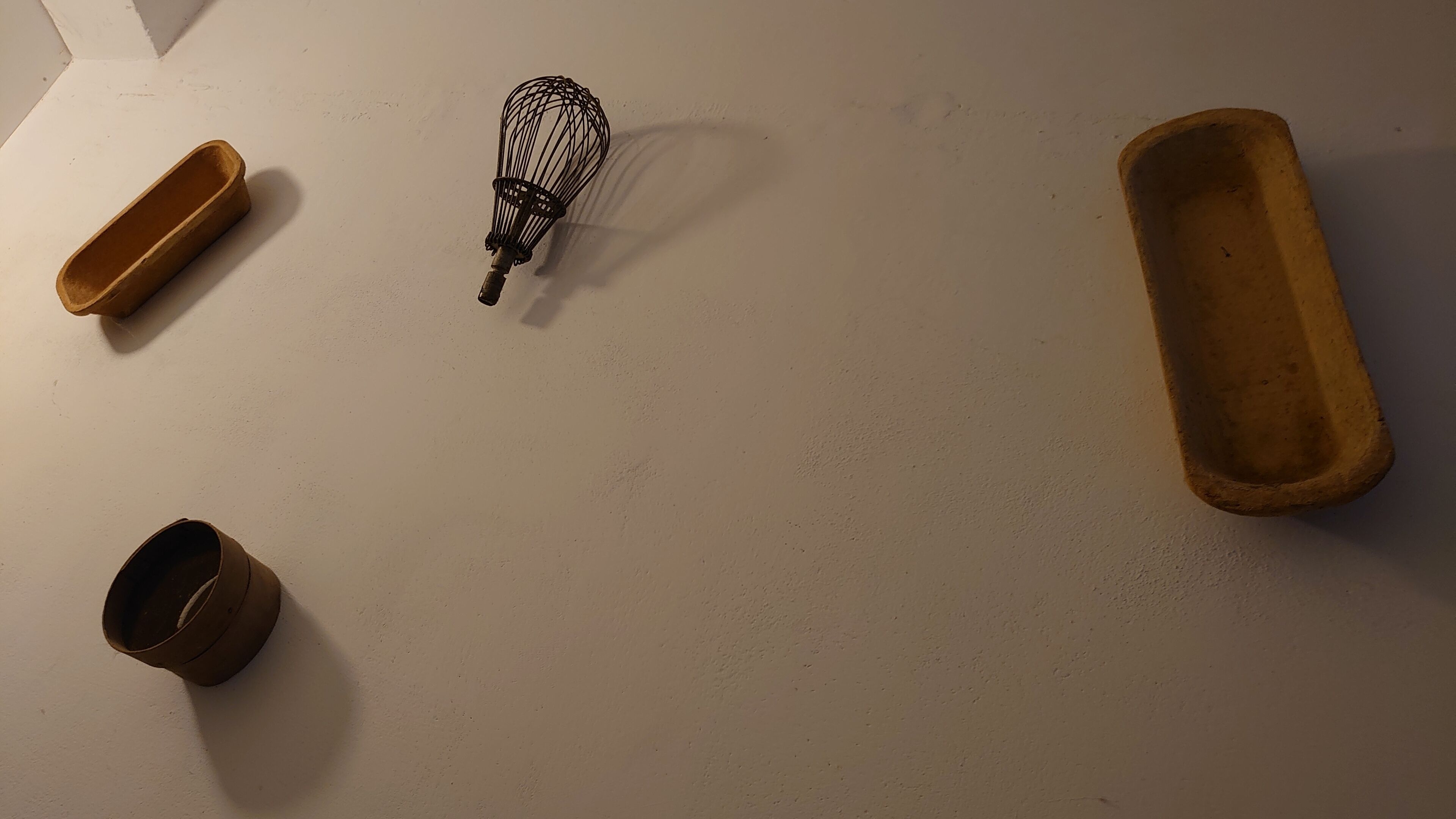 Una habitación con una pared blanca y tres objetos: un batidor, un recipiente marrón pequeño y una bandeja marrón más grande.