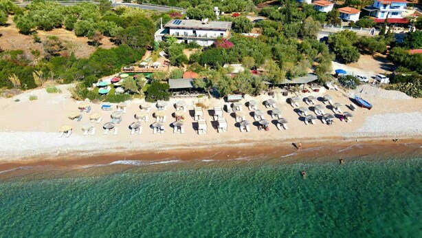 Oversigtsbillede af en strand med liggestole og parasoller, et klart blåt hav og bygninger i baggrunden.
