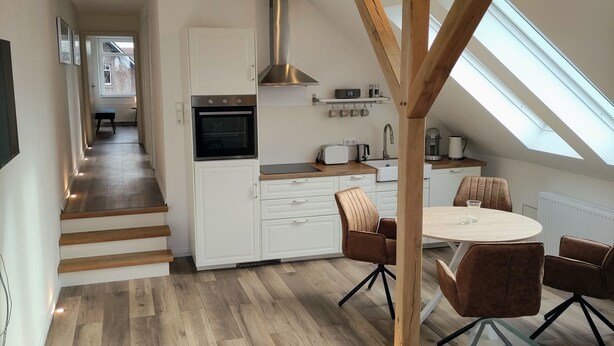 A modern kitchen with white cabinets, a built-in oven, and a stainless steel range hood. There is a dining area with a round table and brown chairs. The floor is wooden, and there are skylights above the kitchen. - Haus im Seewind IV, 70 sqm - country house with large garden, near Friesenstrand (Butjadingen)