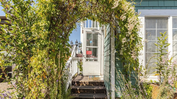 Un escalier en bois mène à une porte blanche dotée d’une fenêtre, le tout étant entouré d’une végétation luxuriante et de plantes en fleurs.