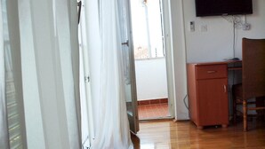 A room with wooden flooring, a desk with a chair, a television mounted on the wall, and a door leading to an outdoor area.