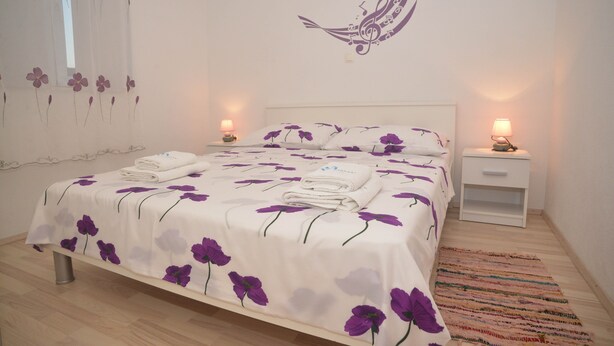 Un lit bien fait, recouvert d’une couvre-lit à motifs floraux, deux tables de chevet avec des lampes, un mur orné de motifs floraux et une fenêtre avec un rideau léger.