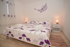 A neatly made bed with a floral bedspread, two bedside tables with lamps, a wall with a floral design, and a window with a sheer curtain. - Barić- Standard One bedroom Apartment with Balcony (A3) (Biograd na Moru)