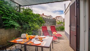 Eine Terrassensitzgruppe im Freien mit Tisch für zwei Personen, umgeben von roten Stühlen, einer Steinmauer und Blick auf benachbarte Gebäude.