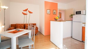 Eine kompakte Küche mit einem weißen Kühlschrank, einer Mikrowelle und einer kleinen Kochinsel. Es gibt einen Essbereich mit einem weißen Tisch und Stühlen, sowie ein orangefarbenes Sofa mit gemusterten Kissen.