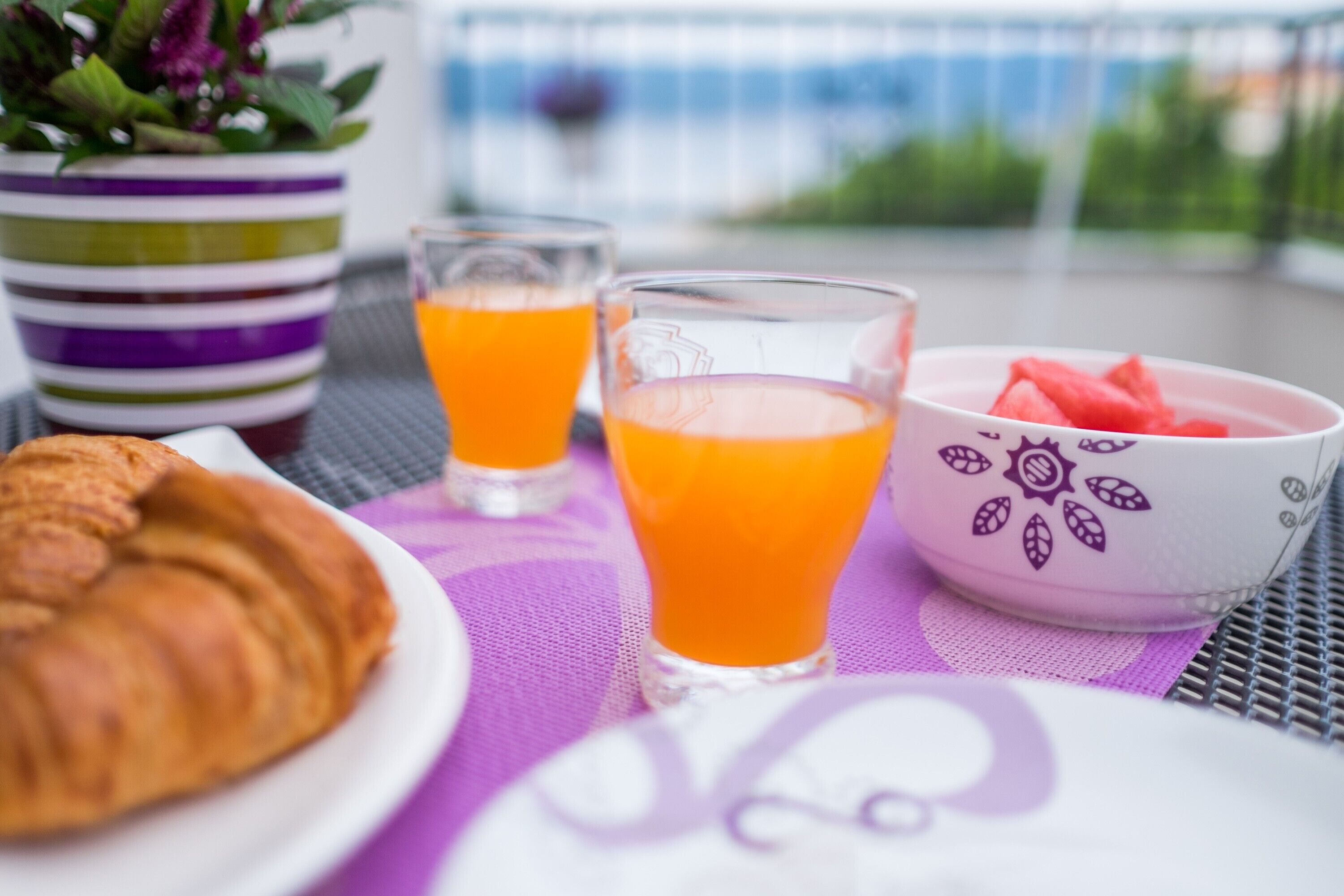 Una escena de desayuno con un croissant, dos vasos de zumo de naranja, un cuenco de sandía y una planta en maceta a rayas.