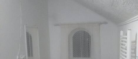 Una habitación pequeña y funcional con un escritorio blanco, un taburete negro, un cubo de basura pequeño y un radiador blanco.