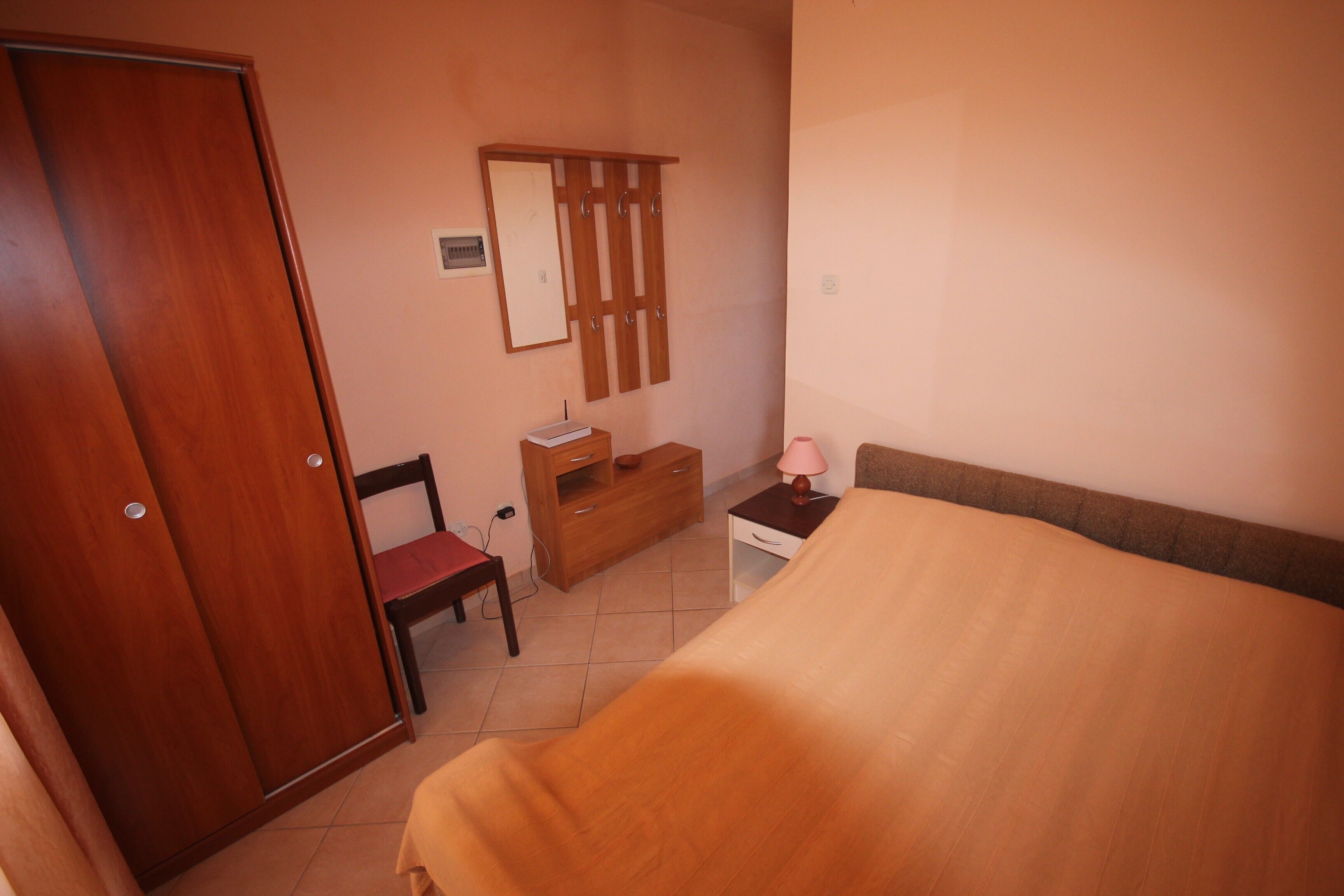 A bedroom with a bed, a wooden wardrobe, a bedside table with a lamp, a chair, and a dresser.