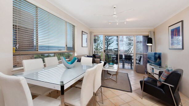 A modern living room with a glass dining table, white chairs, a black sofa, a television, and a balcony with outdoor furniture. - Kookaburra Court U6 | Panoramic Ocean Views by Beach (Caloundra)