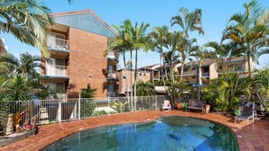 A hotel with a swimming pool surrounded by palm trees and a fenced area. - Kookaburra Court U6 | Panoramic Ocean Views by Beach (Caloundra)