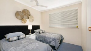 A bedroom with two beds, a ceiling fan, wall art, and a window with blinds. - Kookaburra Court U6 | Panoramic Ocean Views by Beach (Caloundra)
