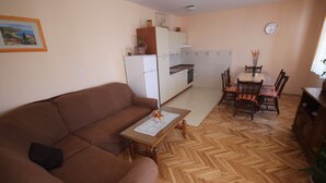 A living room with a brown sofa, a wooden coffee table, and a dining area with a table and chairs. A kitchen with white cabinets and appliances is visible in the background. - Njaco - Three Bedroom Apartment with Sea View (Razanac)