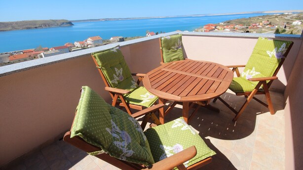 Un balcón con una mesa de madera y cuatro sillas, que ofrece vistas al mar y a casas a lo largo de la costa.