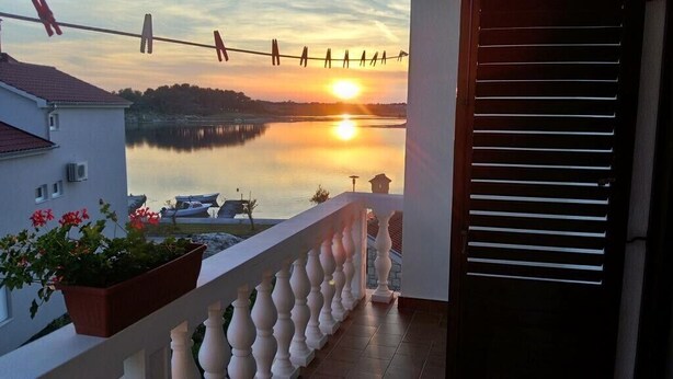 Een balkon met uitzicht op een meer bij zonsondergang, een rij boten en een potplant met rode bloemen.
