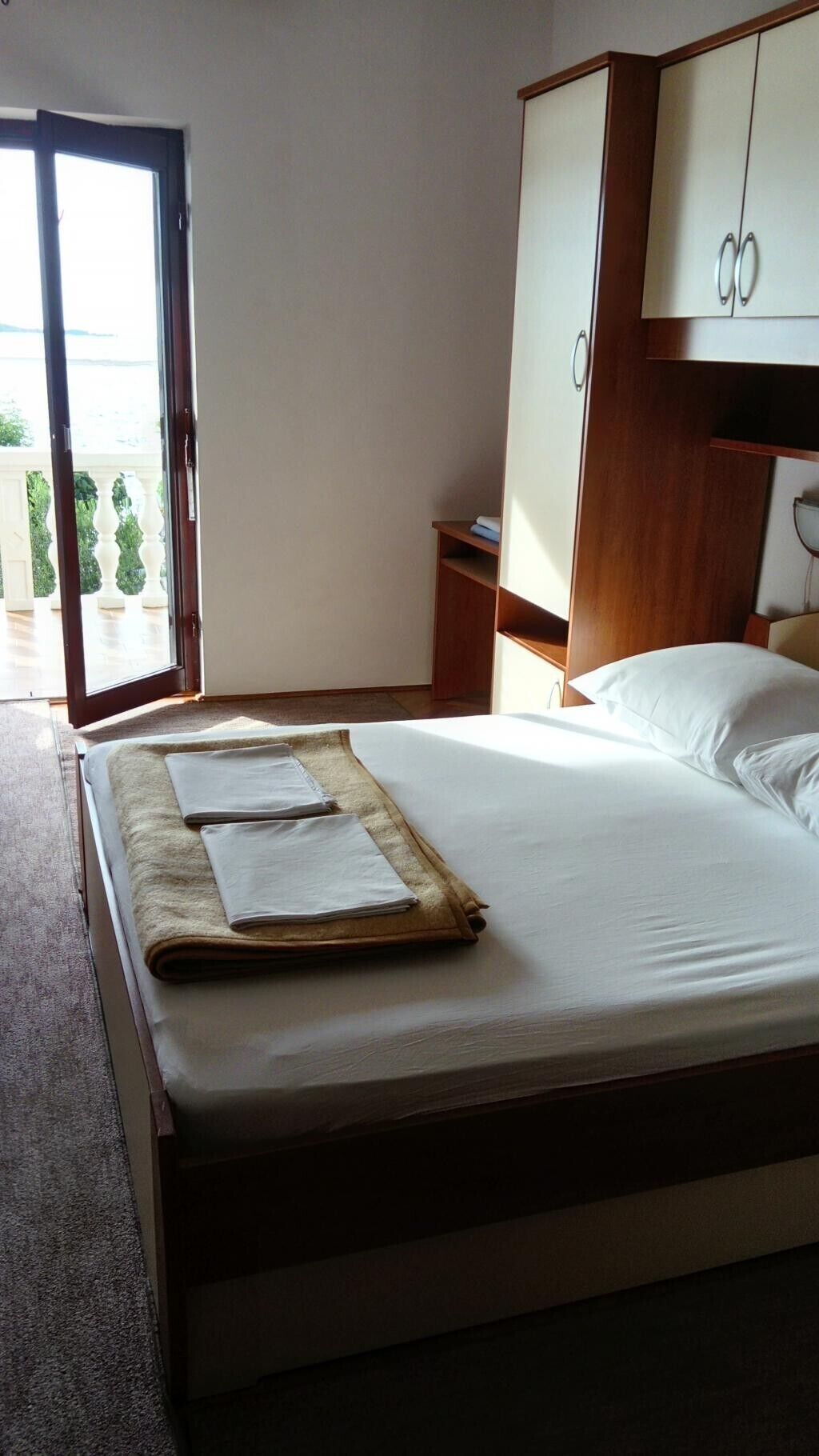 A neatly made bed with a folded blanket and pillows, a wooden wardrobe, and a view of the outdoors through a glass door.