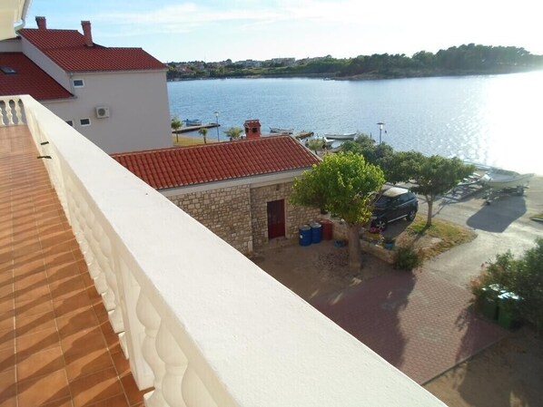 A balcony with a view of a lake, a stone building with a red roof, and a parked car.