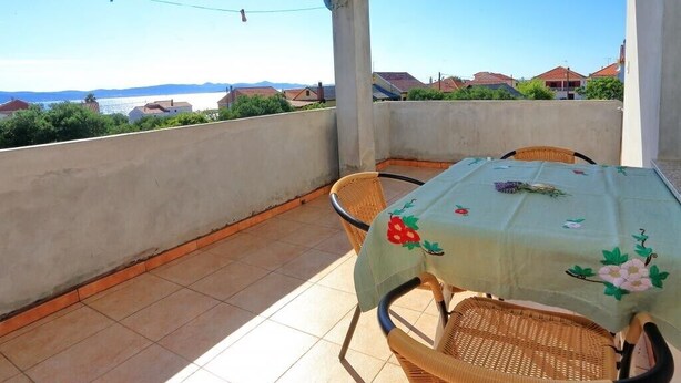 A balcony with a table covered by a floral tablecloth, wicker chairs, and a view of houses and water in the background. - Apartments Suzana - Studio Apartment with Sea View (Bibinje)
