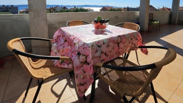 Ein Tisch mit einer blumigen Tischdecke und einer Obstschüssel auf einem Balkon mit Korbstühlen.