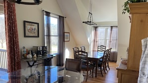 A dining area with a glass table and wooden chairs, a coffee station, and a view of the outdoors through large windows.