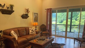 A living room with a brown leather sofa, a glass coffee table, and a wooden floor. There is a balcony with white chairs and a view of greenery.