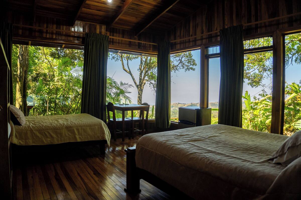 A room with two beds, a table, and a chair, with large windows offering a view of lush greenery.