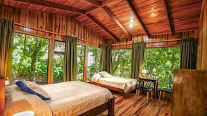 A wooden cabin with two beds, a table, and large windows overlooking a forest. - Hotel Flor de Bromelia (Monteverde)