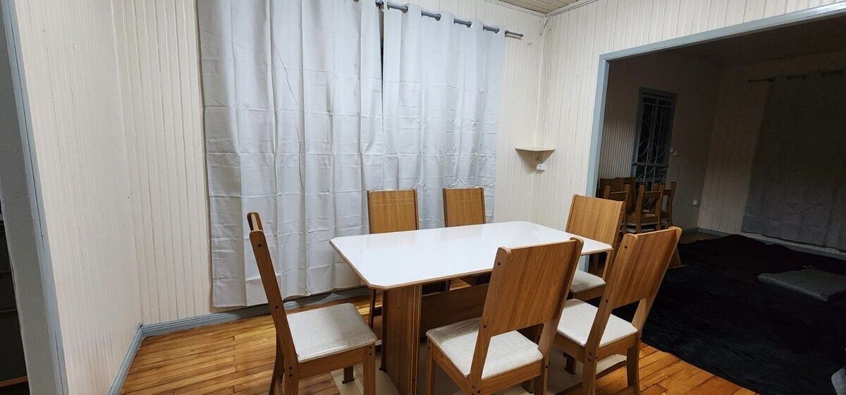 Un comedor con una mesa blanca y sillas de madera, una cortina blanca y un estante pequeño.