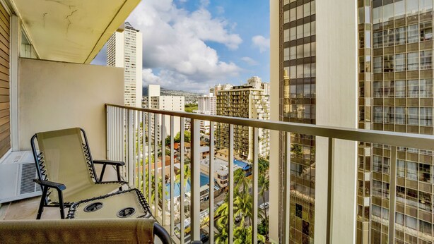 A balcony with a chair and a table, overlooking a cityscape with tall buildings and palm trees. - Marine Surf by Luana HI Vacation Rental (Honolulu)
