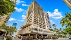 A tall hotel with a modern design, featuring a prominent entrance and a covered area with stairs leading up to it. - Marine Surf by Luana HI Vacation Rental (Honolulu)
