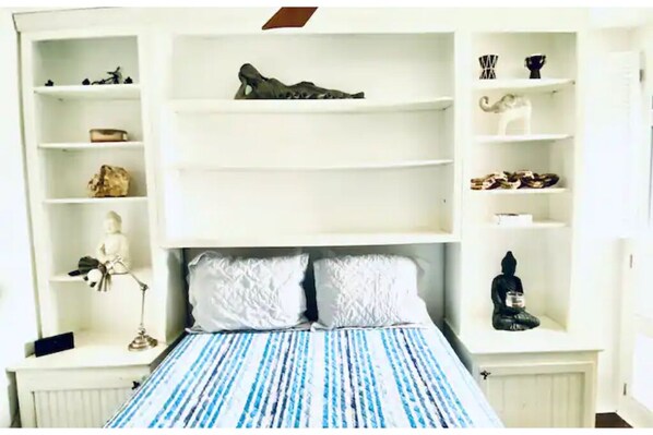 Une chambre à coucher avec un lit, deux oreillers, une table de chevet, une lampe et une étagère ornée d’objets décoratifs.