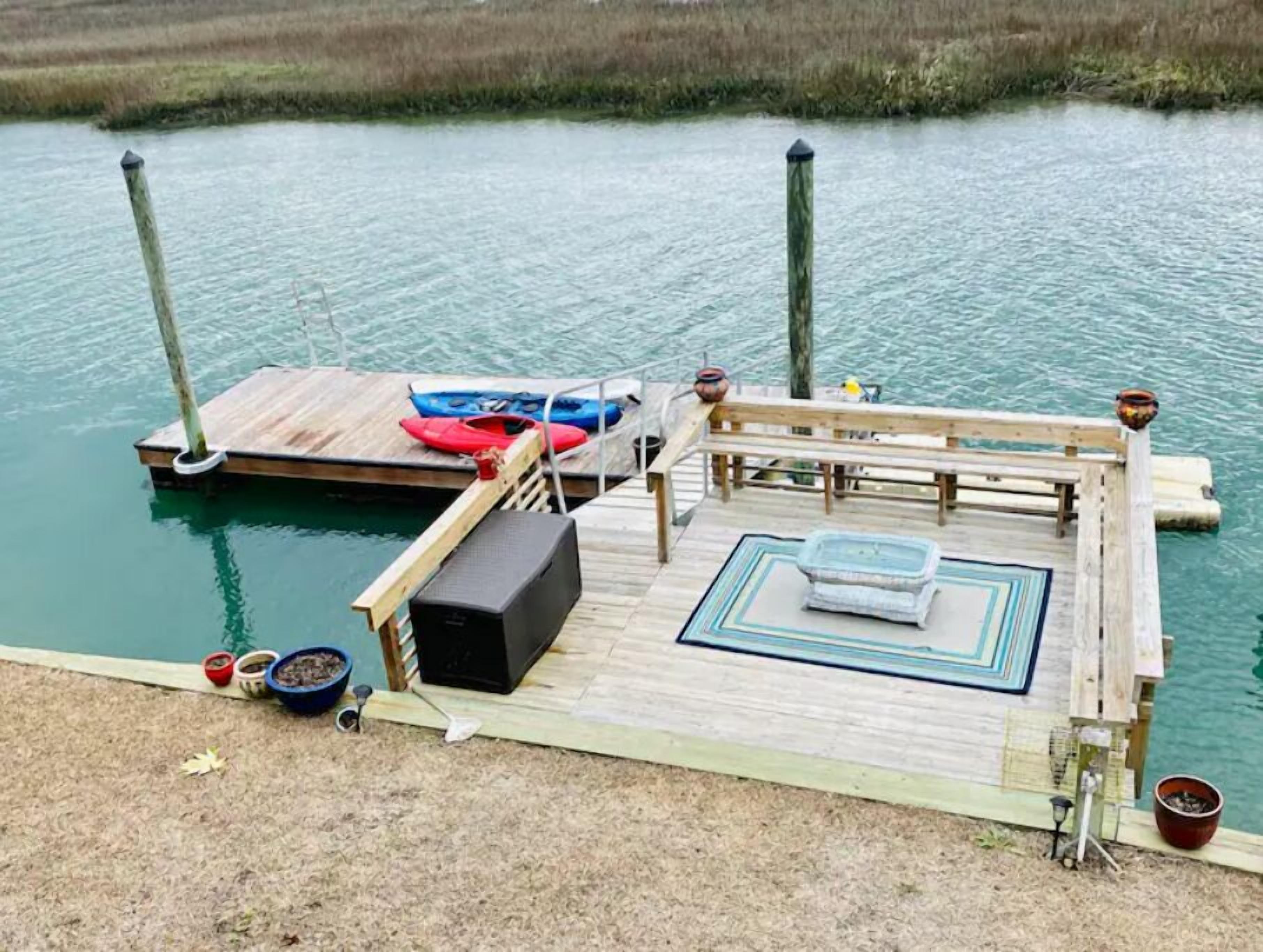 A wooden dock with a bench, a small table, and a speaker. There are kayaks and a paddleboard on the dock.