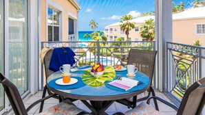Une table ronde recouverte d’une nappe bleue, entourée de chaises en osier, sur un balcon d’où l’on peut admirer un bâtiment et des palmiers.