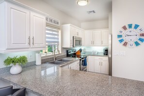 A kitchen with white cabinets, a granite countertop, a stainless steel oven, a clock, and a sign that says 'AT THE OCEAN'. - Famous 7-Mile Beach Retreat - Stunning Views & Summer Savings! (Seven Mile Beach)