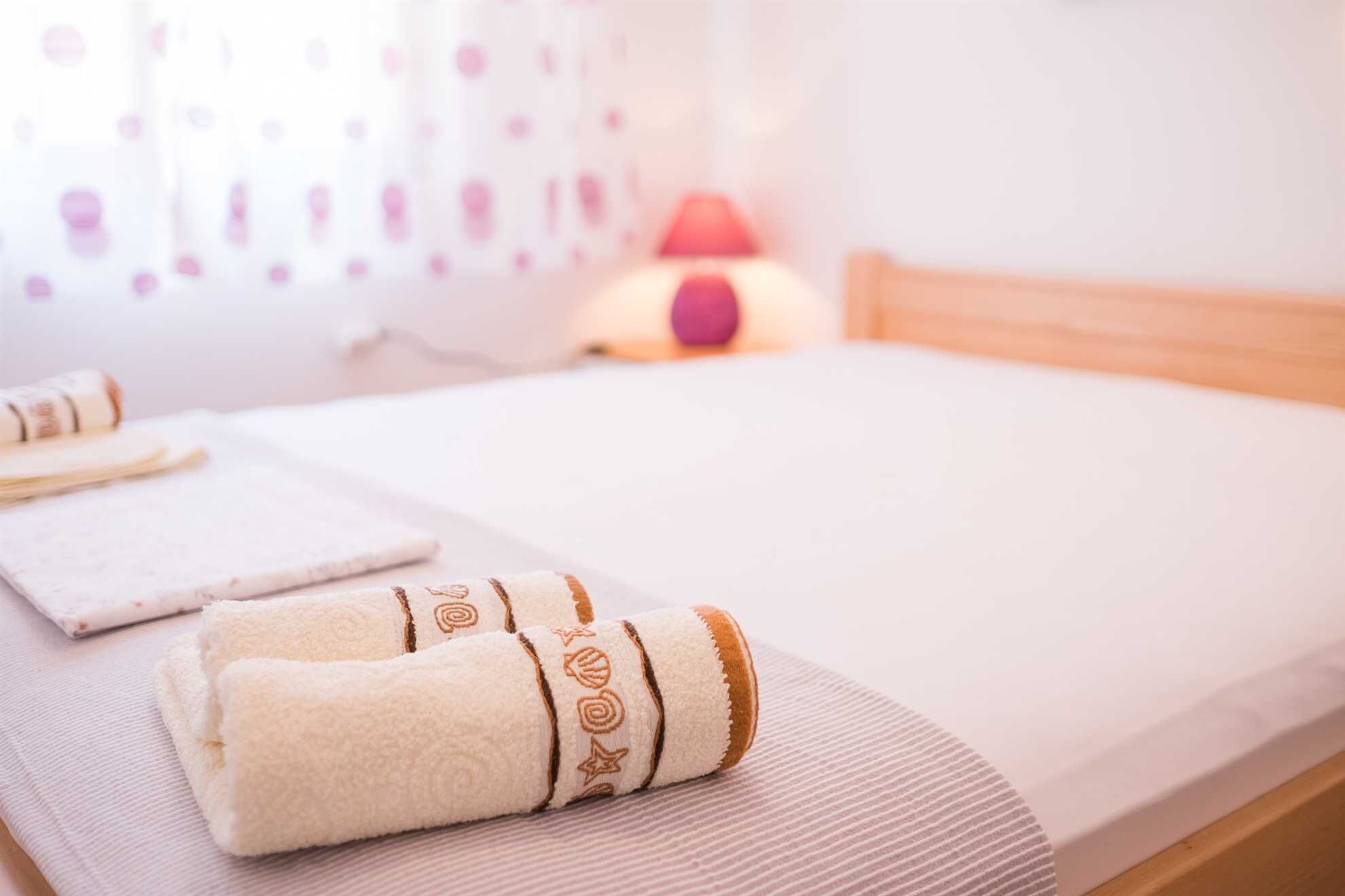A neatly made bed with a white mattress and a white blanket. There are two rolled towels with brown patterns on the bed. A red lamp is on the nightstand beside the bed.