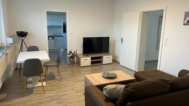 A modern living room with a flat-screen TV, a brown sofa, a wooden coffee table, and a dining area with a white table and two chairs. - A modern renovated apartment with balcony terrace. (Lübbecke)