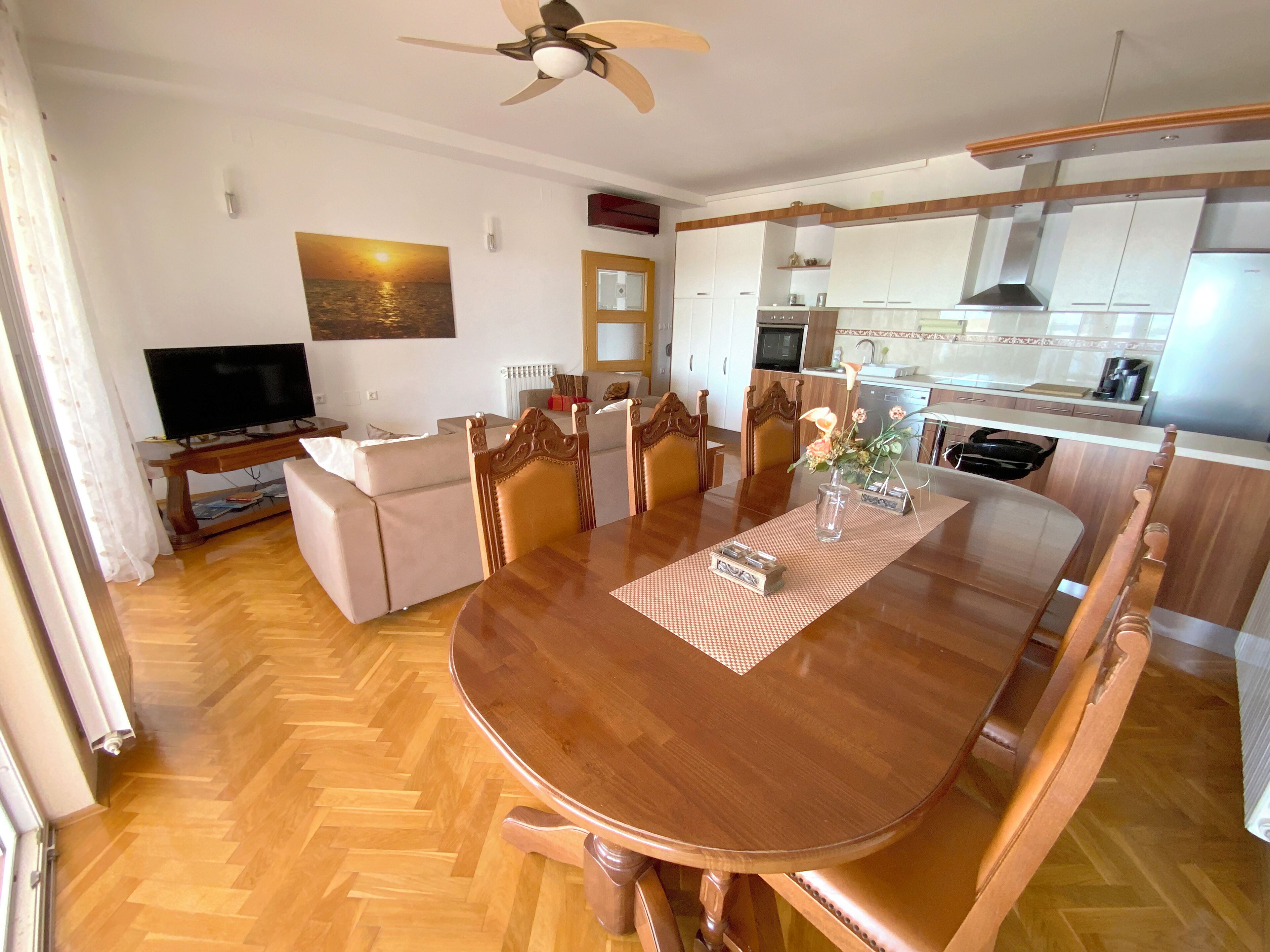 Un comedor con una mesa y sillas de madera, un televisor en la sala de estar y una cocina al fondo.