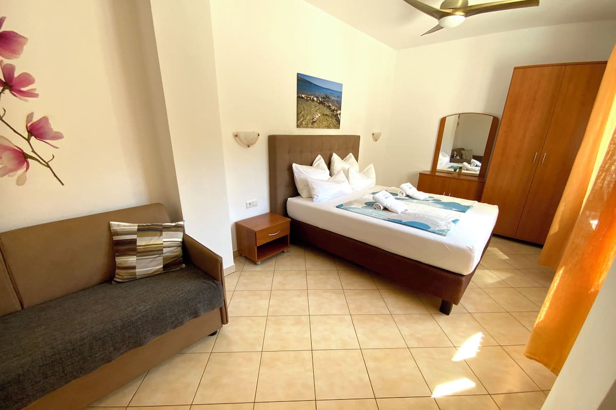 A hotel room with a bed, a nightstand, a sofa, a wooden wardrobe, a ceiling fan, and a wall-mounted picture.