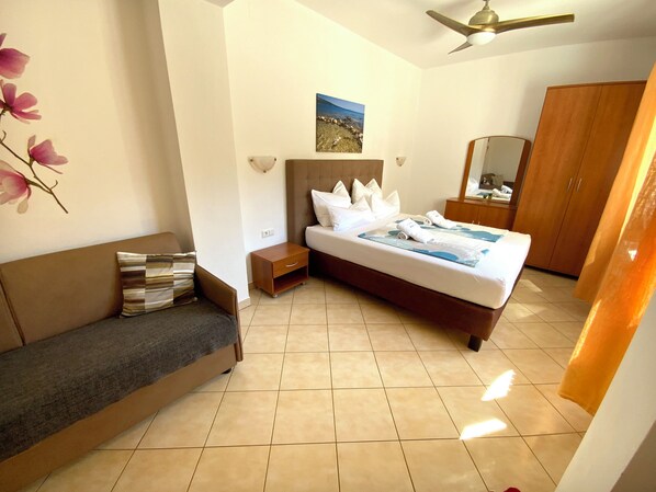Habitación de hotel con cama, mesita de noche, sofá, armario de madera, ventilador de techo e imagen colgada en la pared.