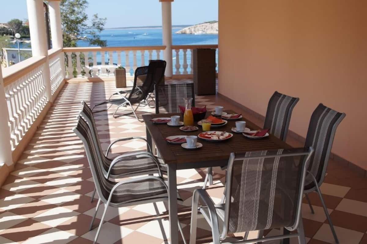 A terrace with a checkered floor, a dining table set with plates, cups, and a bottle, surrounded by chairs.