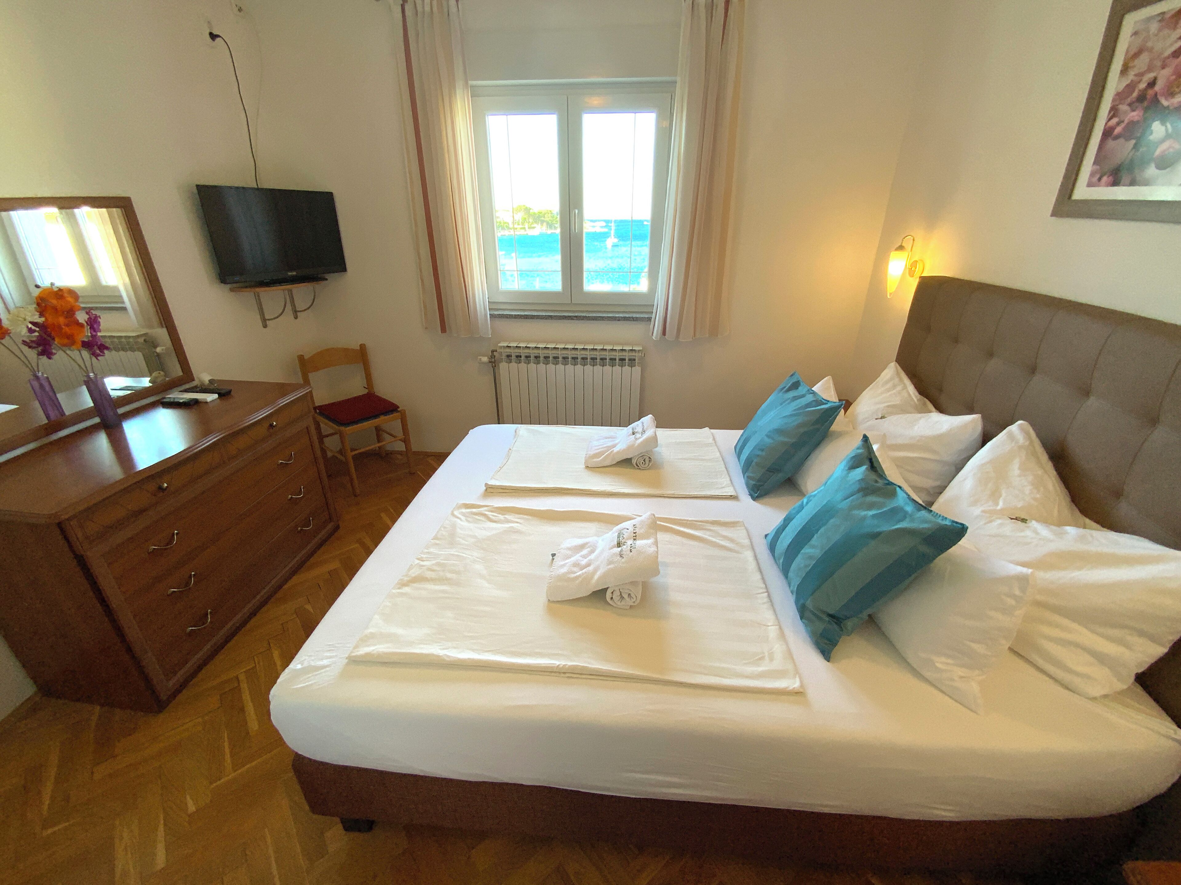 A hotel room with a bed, a dresser, a television, a window with a view, and a radiator.
