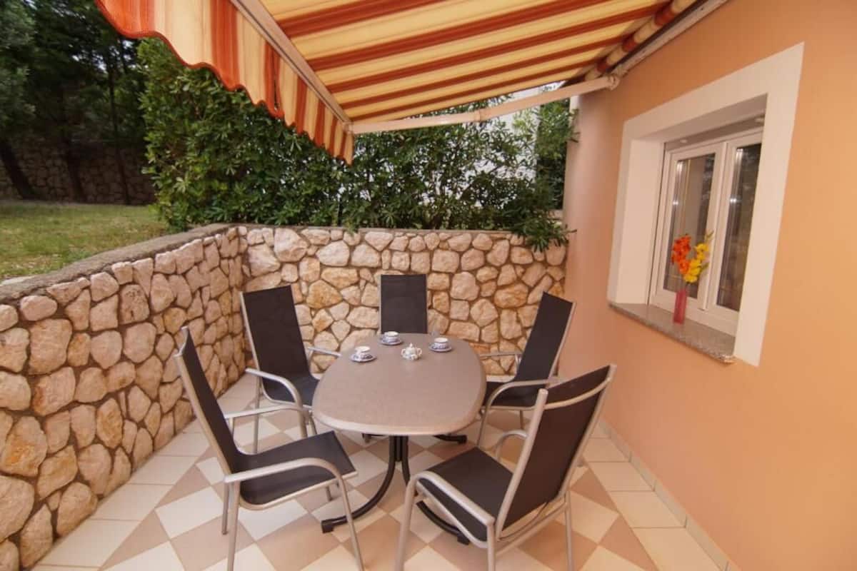 A stone wall patio with a round table and four chairs, an outdoor umbrella, and a window with flowers.