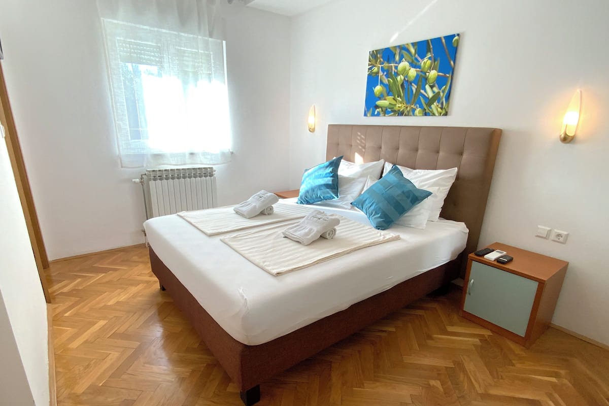 A neatly made bed with white linens and blue pillows, a small bedside table with a phone and remote, and a radiator on the wall.