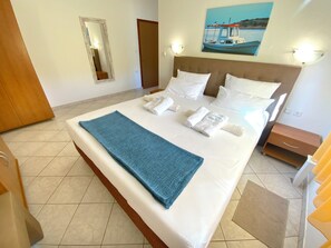 A hotel room with a large bed, white linens, a blue throw blanket, and a painting of a boat on the wall. - Olive House (Pag)