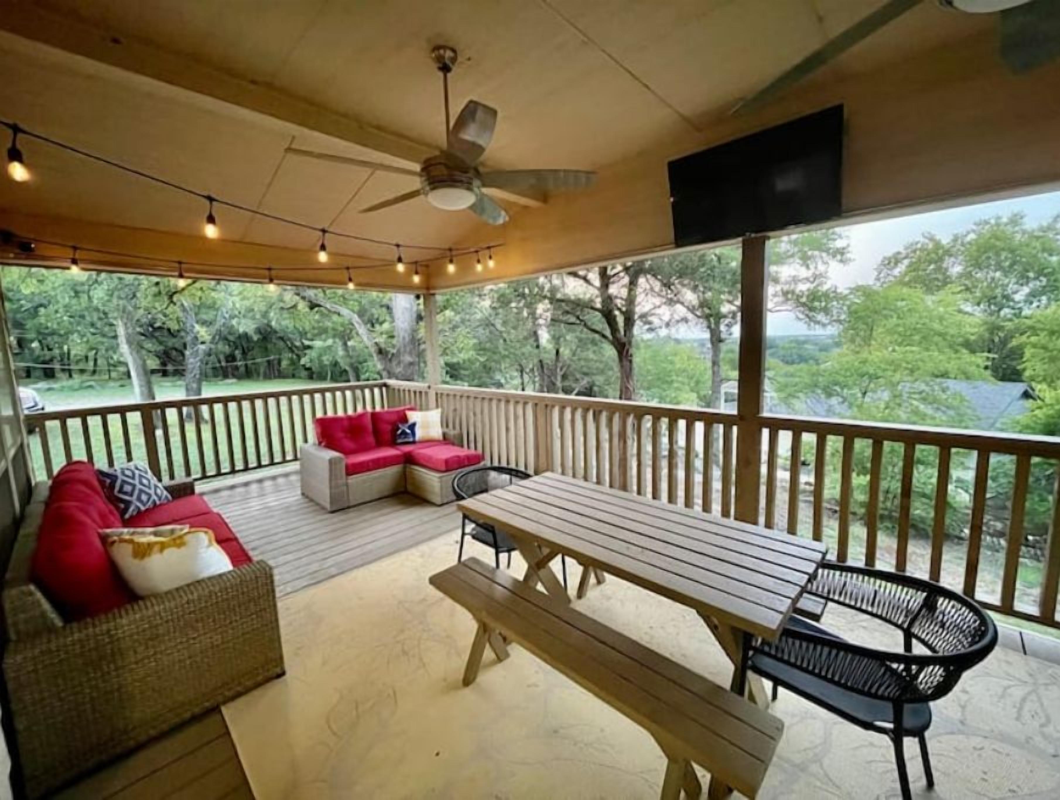 Un espace extérieur abrité doté d’une terrasse en bois, d’un ventilateur de plafond, de guirlandes lumineuses, d’une table en bois, d’un banc et d’un canapé avec des coussins.