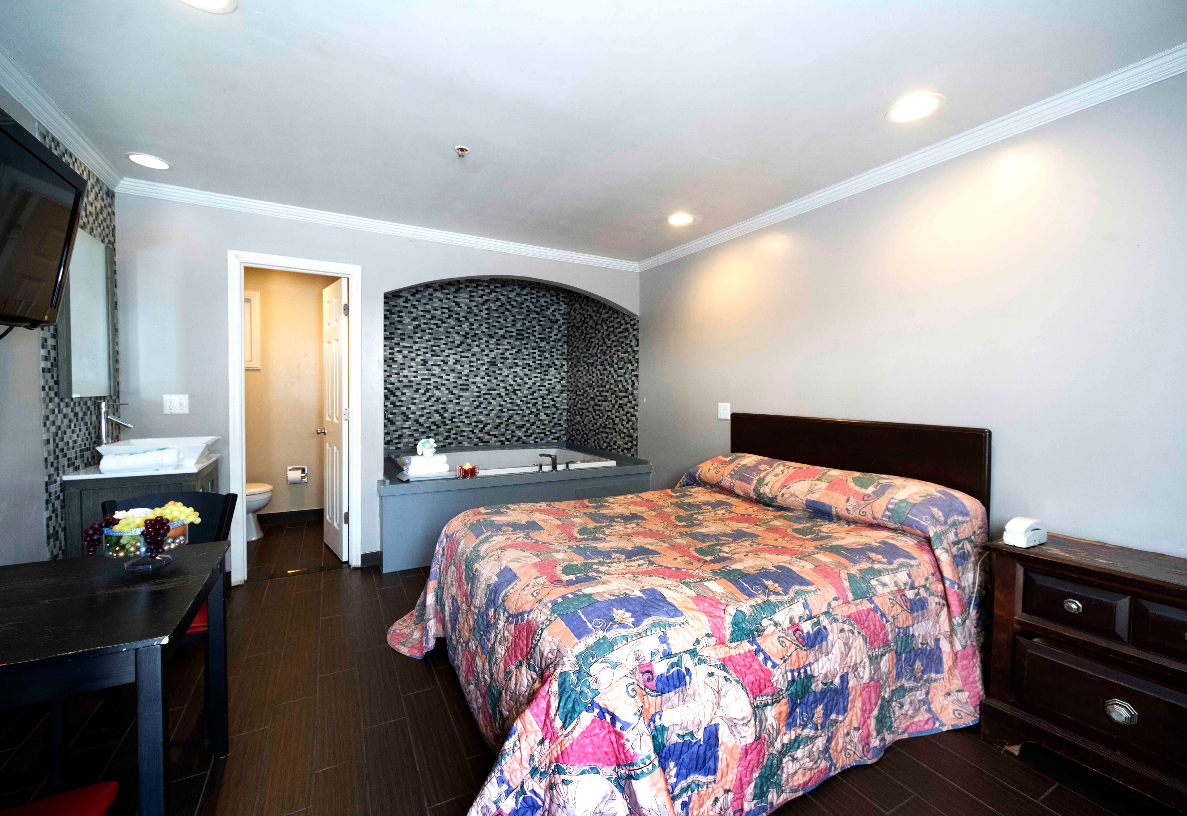 A bedroom with a bed, a desk, a chair, a dresser, a bathroom with a sink and toilet, and a mirror.